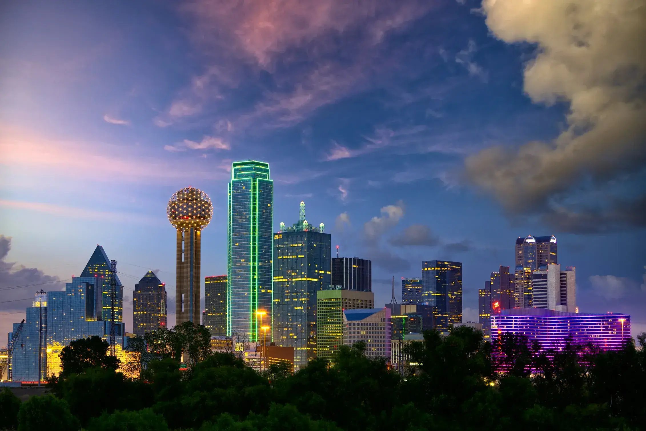 Dallas skyline at dusk featuring Reunion Tower and illuminated downtown skyscrapers beneath dramatic clouds.