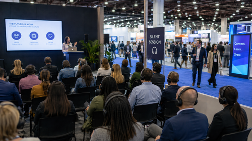 Attendees wearing plain black headsets listen to a silent session presentation inside a busy convention center, with nearby expo activity continuing in the background.