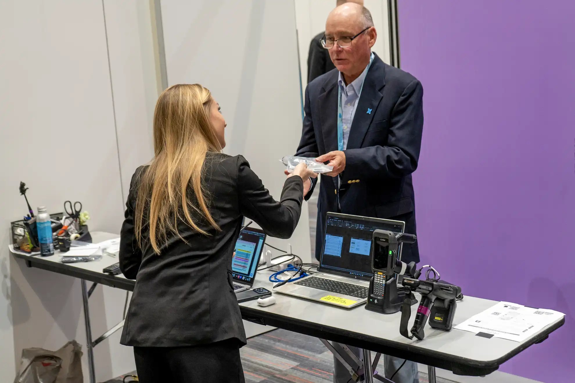 Event staff member hands a wireless listening device to an attendee at an equipment check-in station with laptops and barcode scanners.