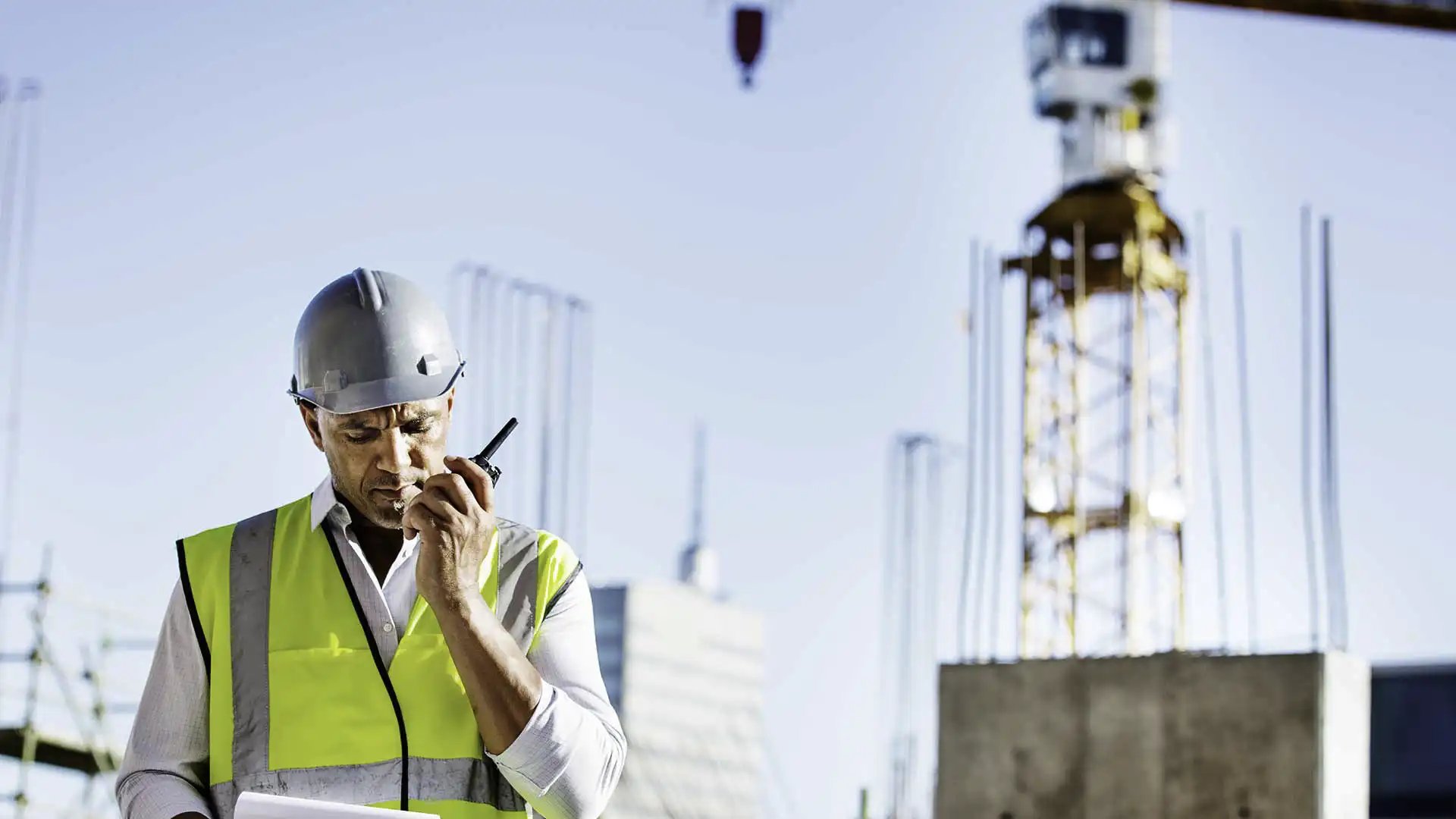 Foreman in hard hat and safety vest speaks into a radio with tower crane in background.