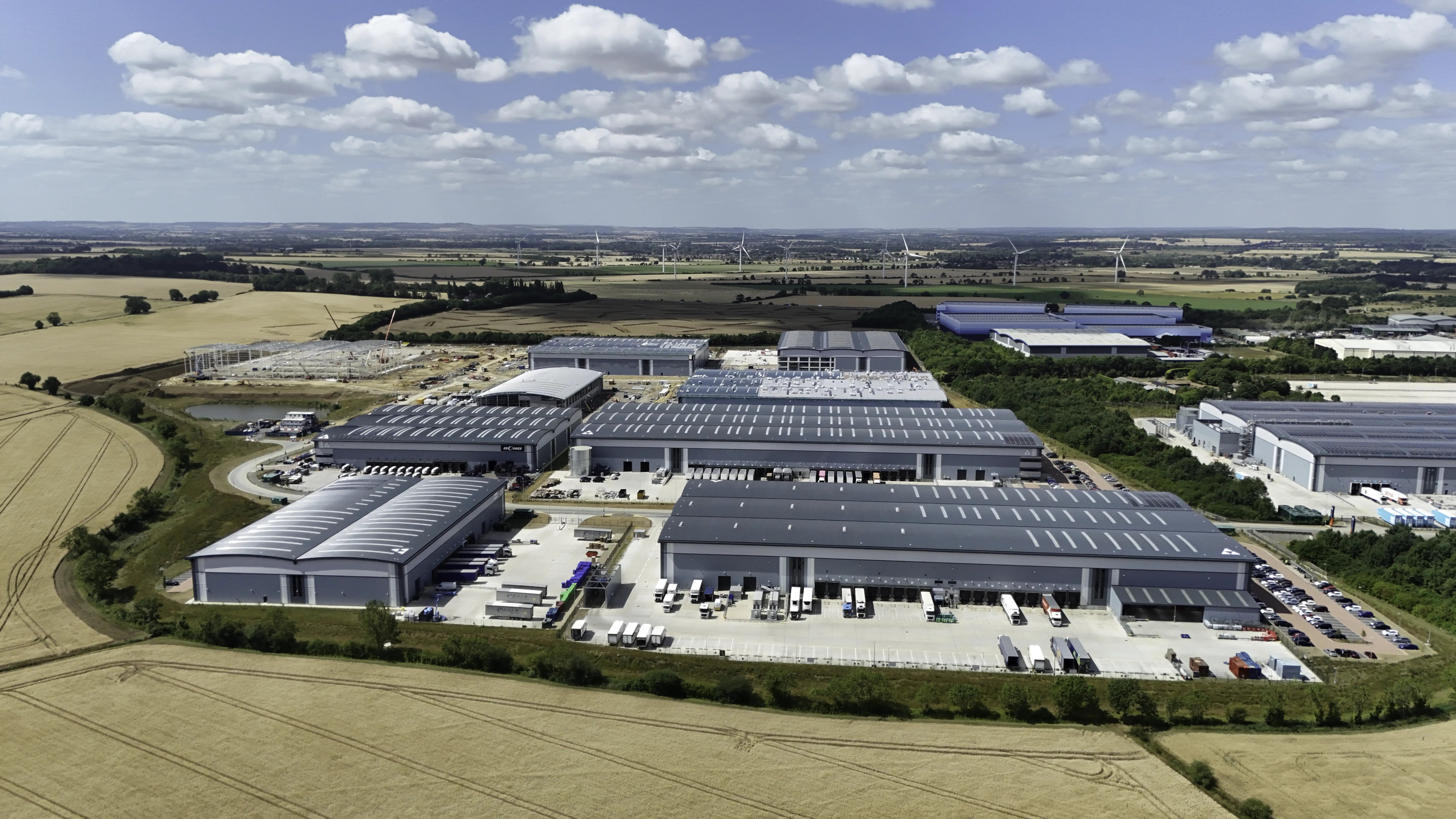 Wide aerial shows multiple industrial buildings, loading docks, and nearby wind turbines.