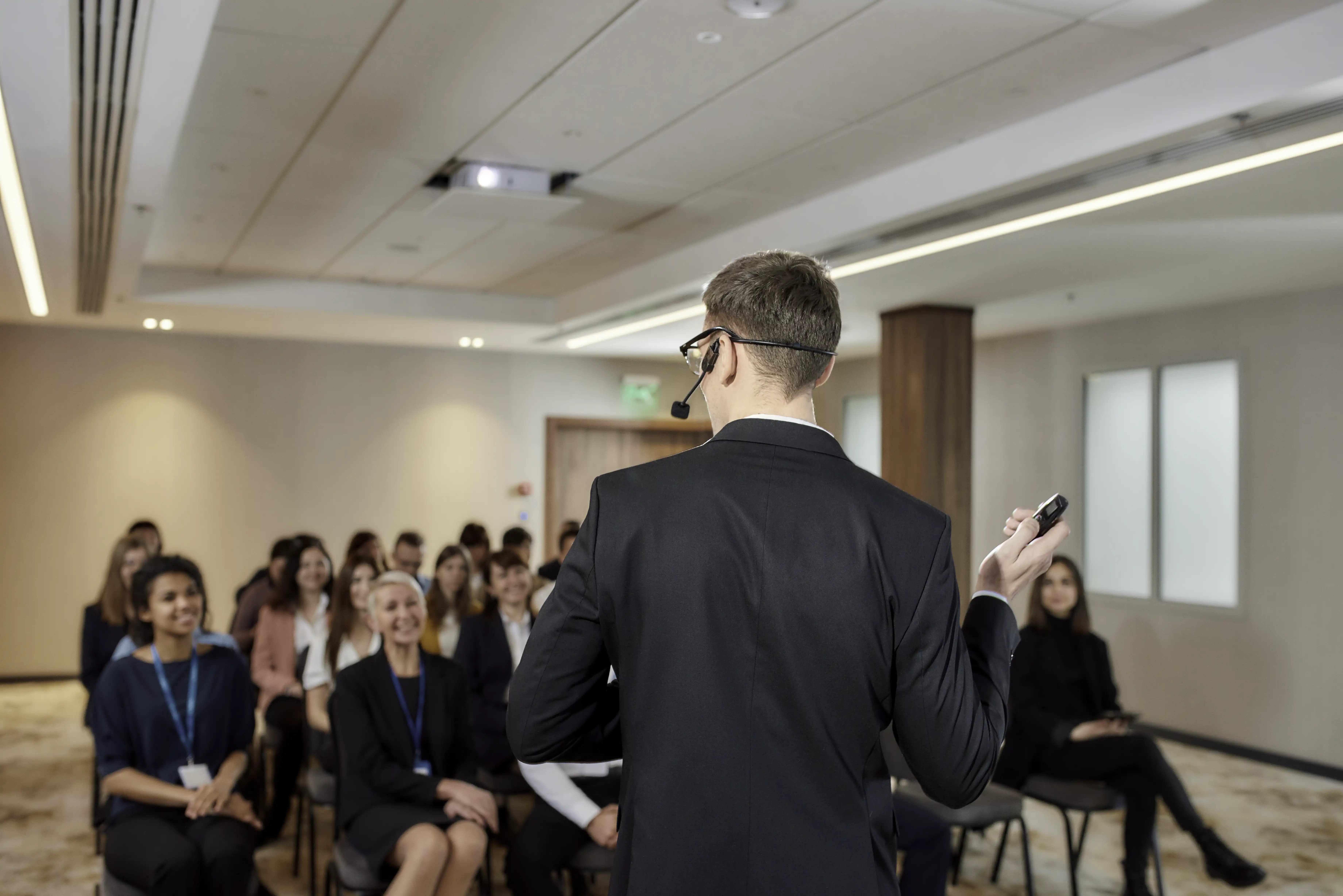 Presenter wearing a headset microphone addresses a seated audience in a conference room.