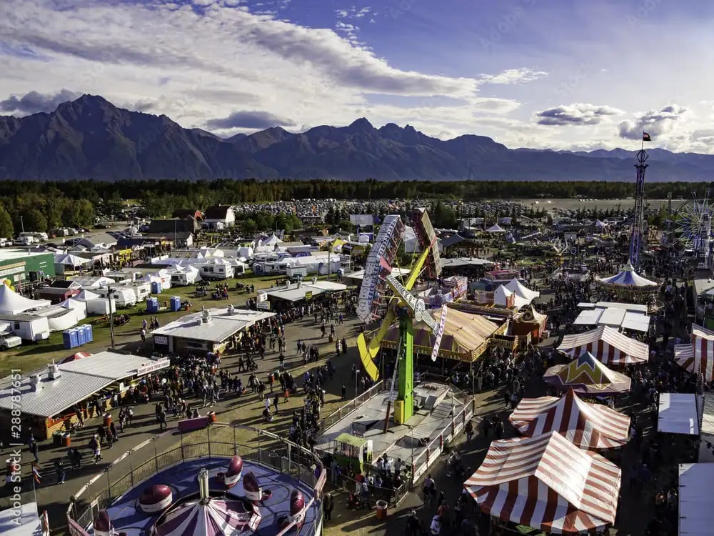 Crowds move through tents and carnival rides at a large fair beneath mountain peaks.