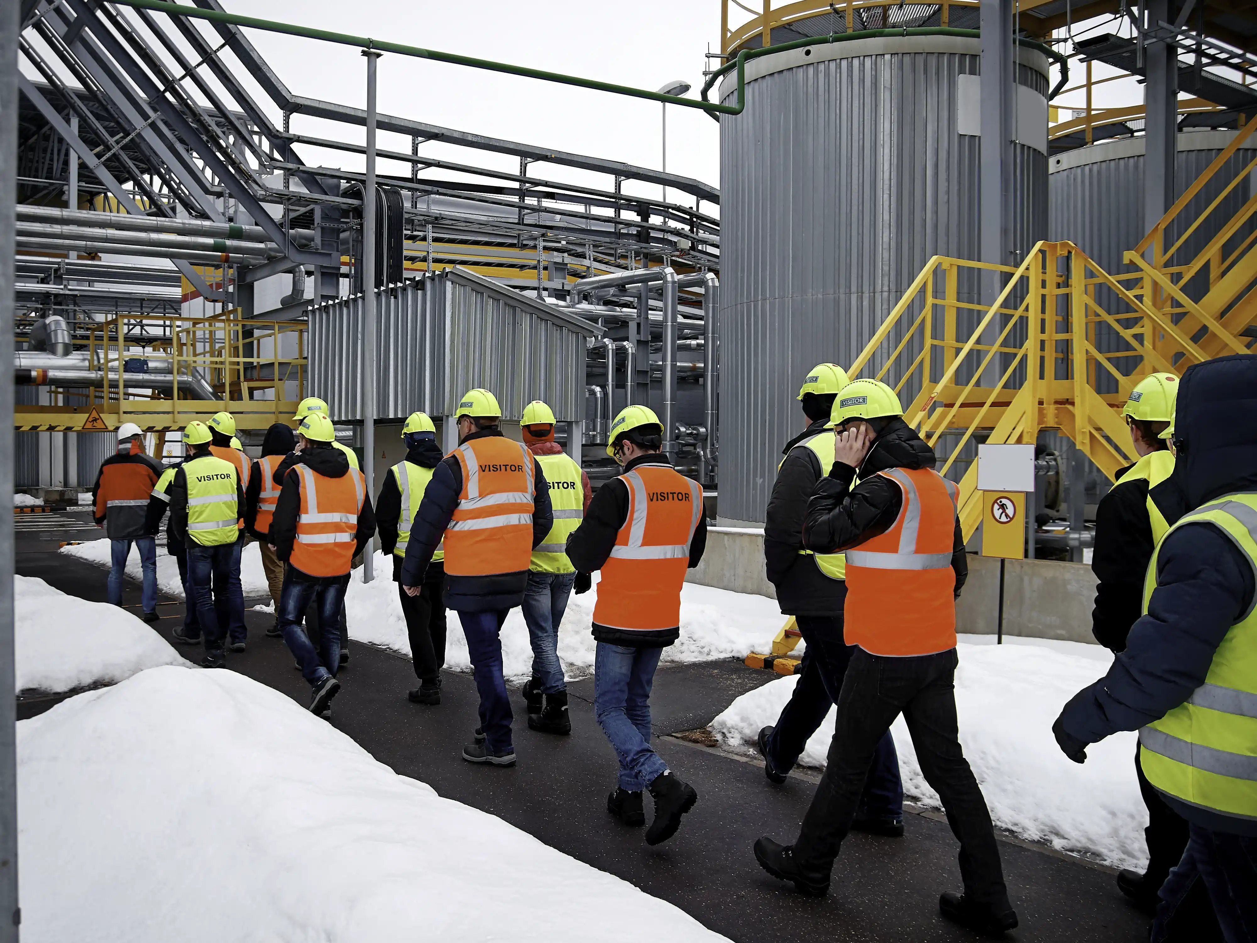 Visitors in high visibility vests and hard hats tour an outdoor industrial site in the snow.