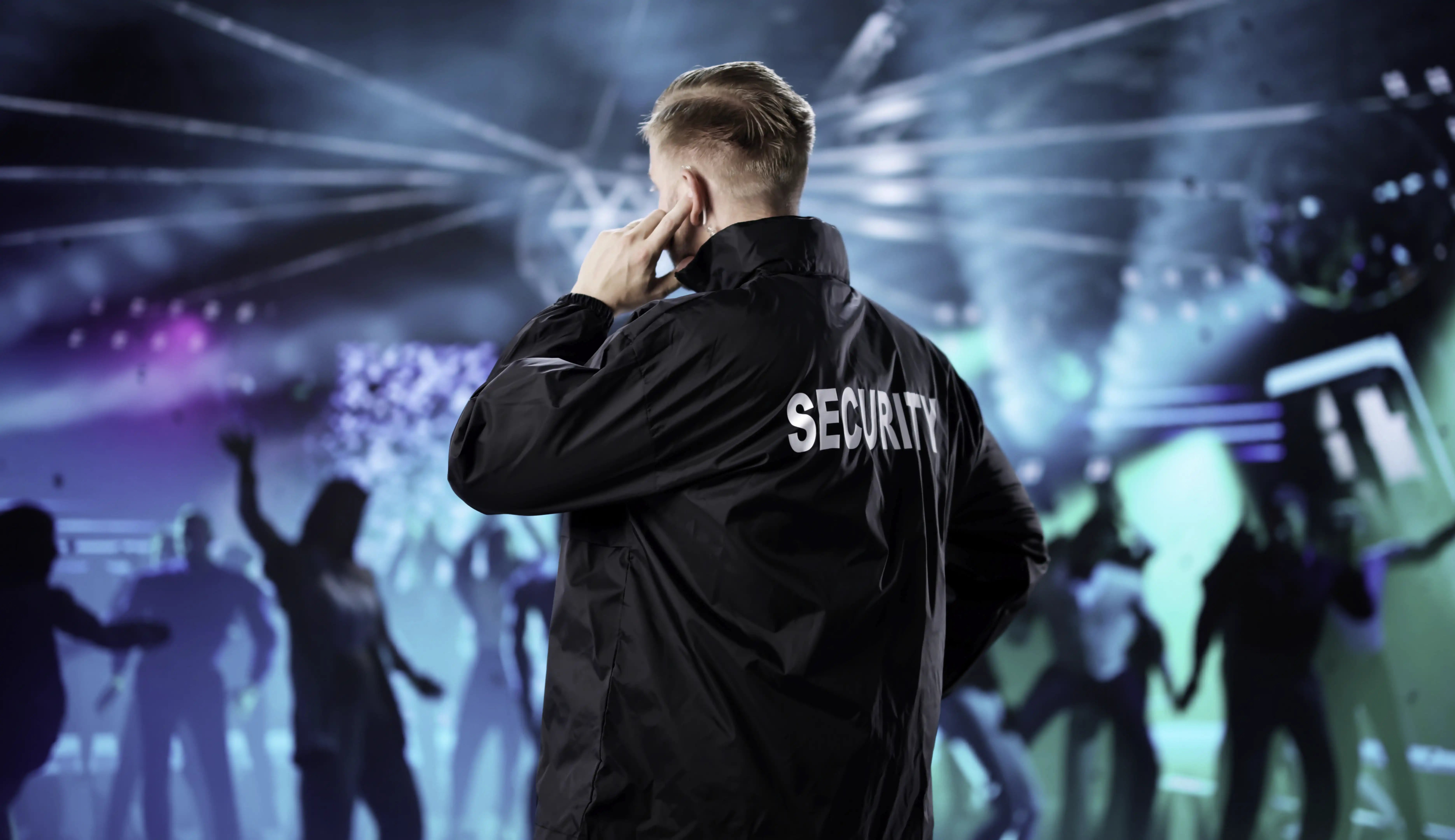 Security staff member watches a lively venue with stage lights and dancing guests.