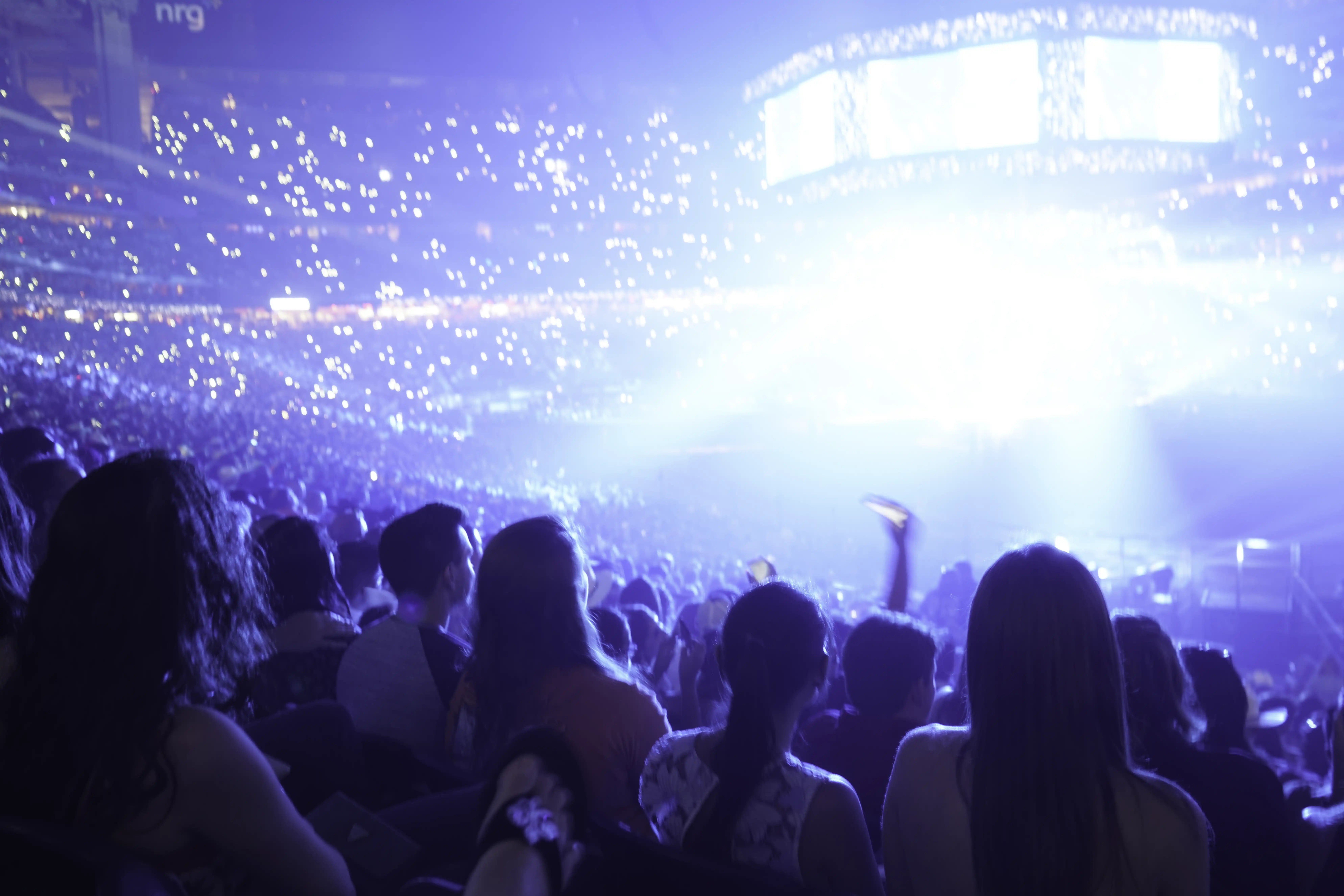 Fans fill a packed indoor arena as stage lights glow and phones light up the crowd.