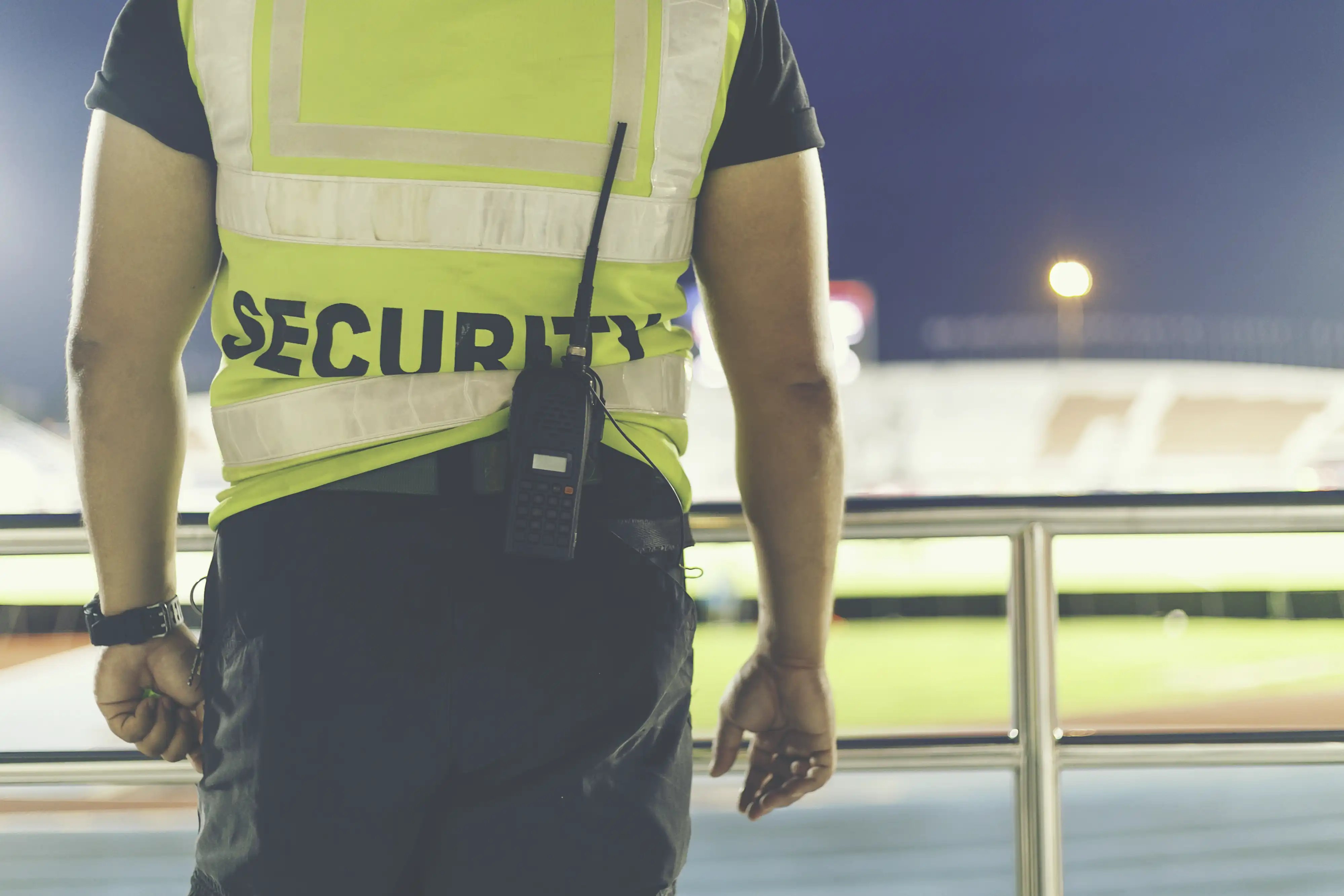Security guard in a high-visibility vest stands by a railing with a handheld two way radio during a stadium event.