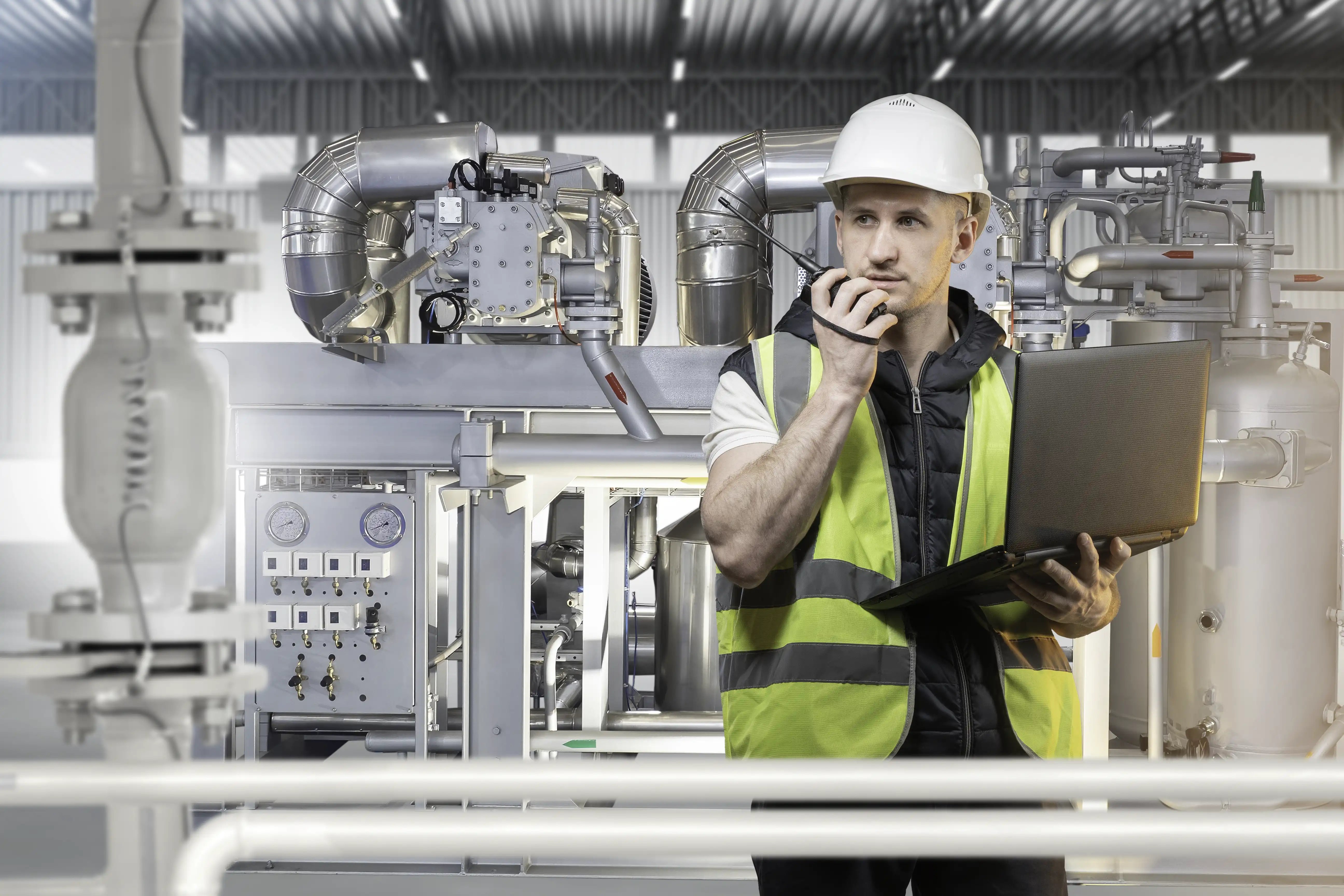 Worker in a hard hat and hi-vis vest uses a two way radio while checking data on a laptop in a processing plant.