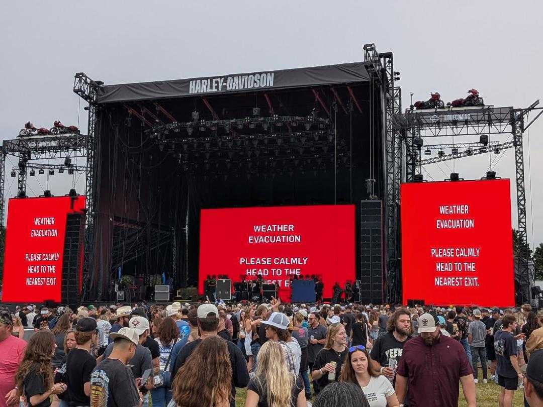 Crowd gathered in front of a large outdoor concert stage labeled “Harley-Davidson,” with three bright red video screens displaying “WEATHER EVACUATION, PLEASE CALMLY HEAD TO THE NEAREST EXIT.”