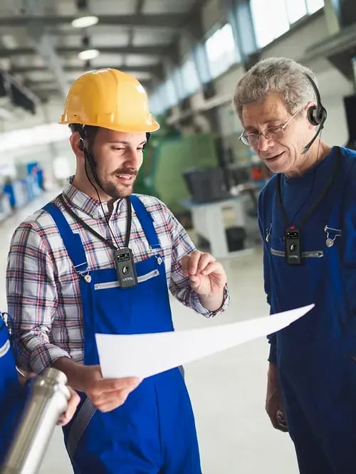 Two workers in overalls looking at plans on a sheet of paper. They are wearing headsets.