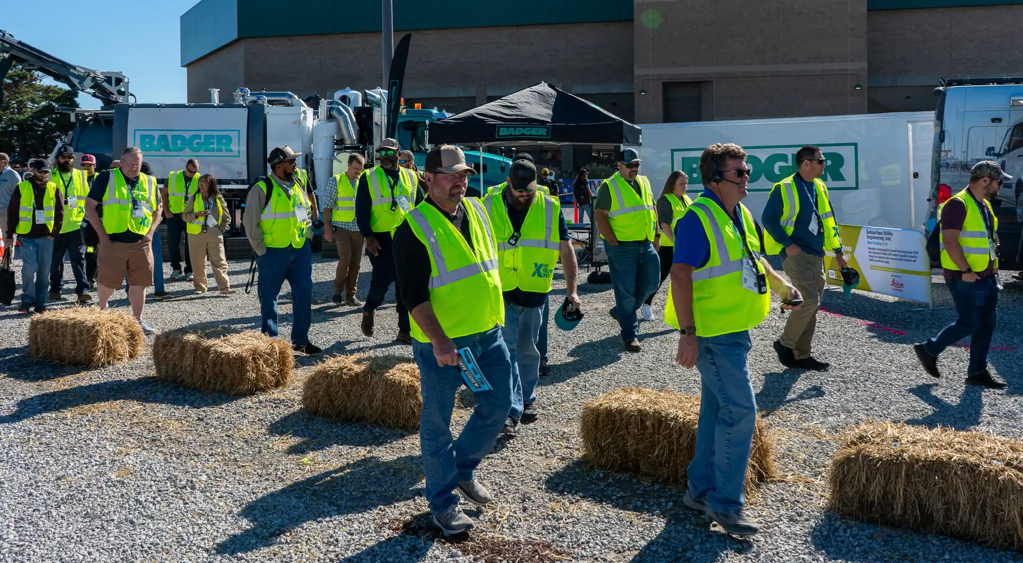 Group of utility professionals wearing high-visibility safety vests walking through an outdoor equipment exhibit at a trade show, using wireless headsets for guided instruction, with heavy machinery and vendor booths in the background.