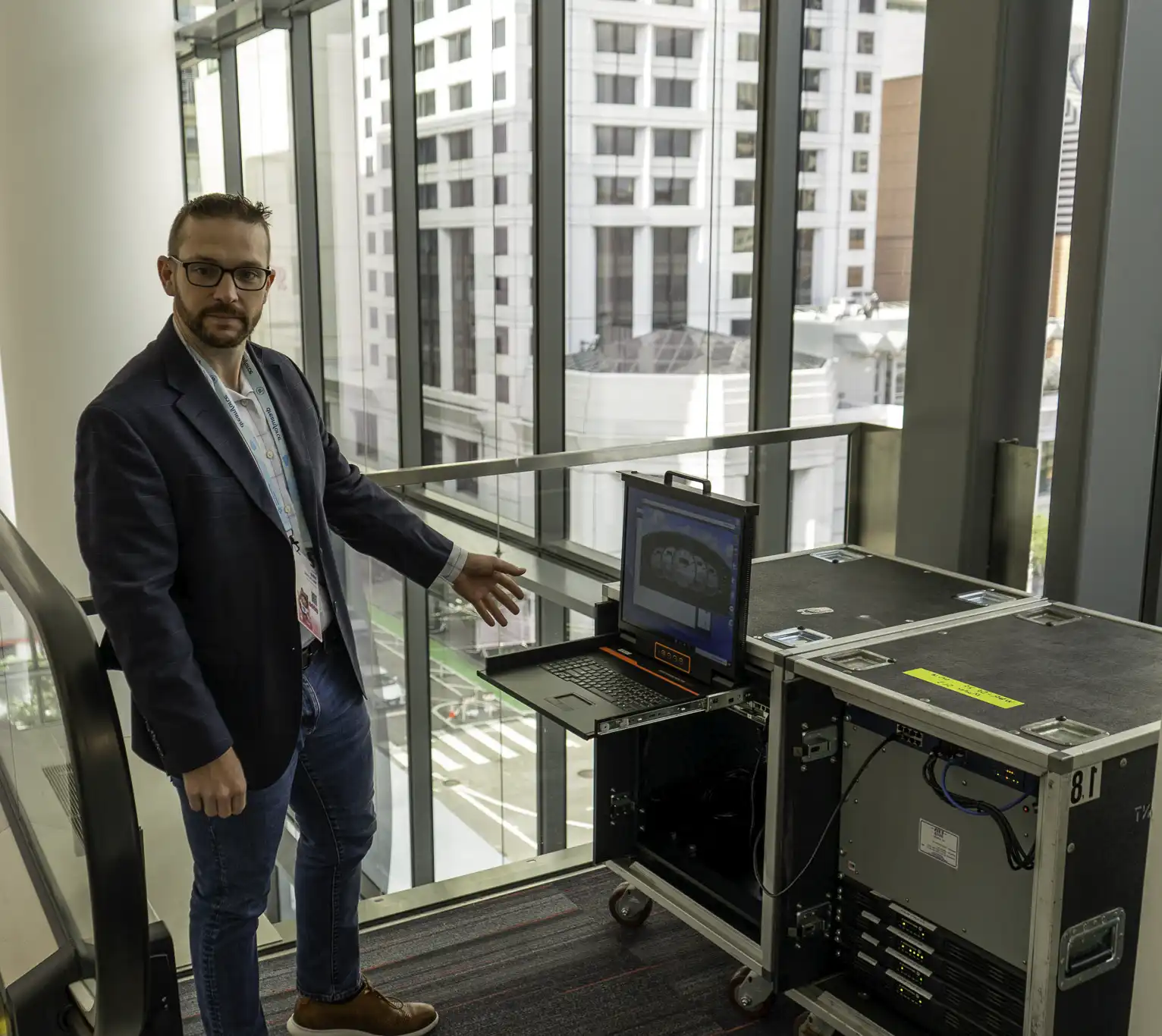 Man wearing glasses and a blazer stands beside a rolling equipment rack with an open laptop, gesturing toward the setup in a glass-walled corridor next to an escalator, with city buildings visible outside.