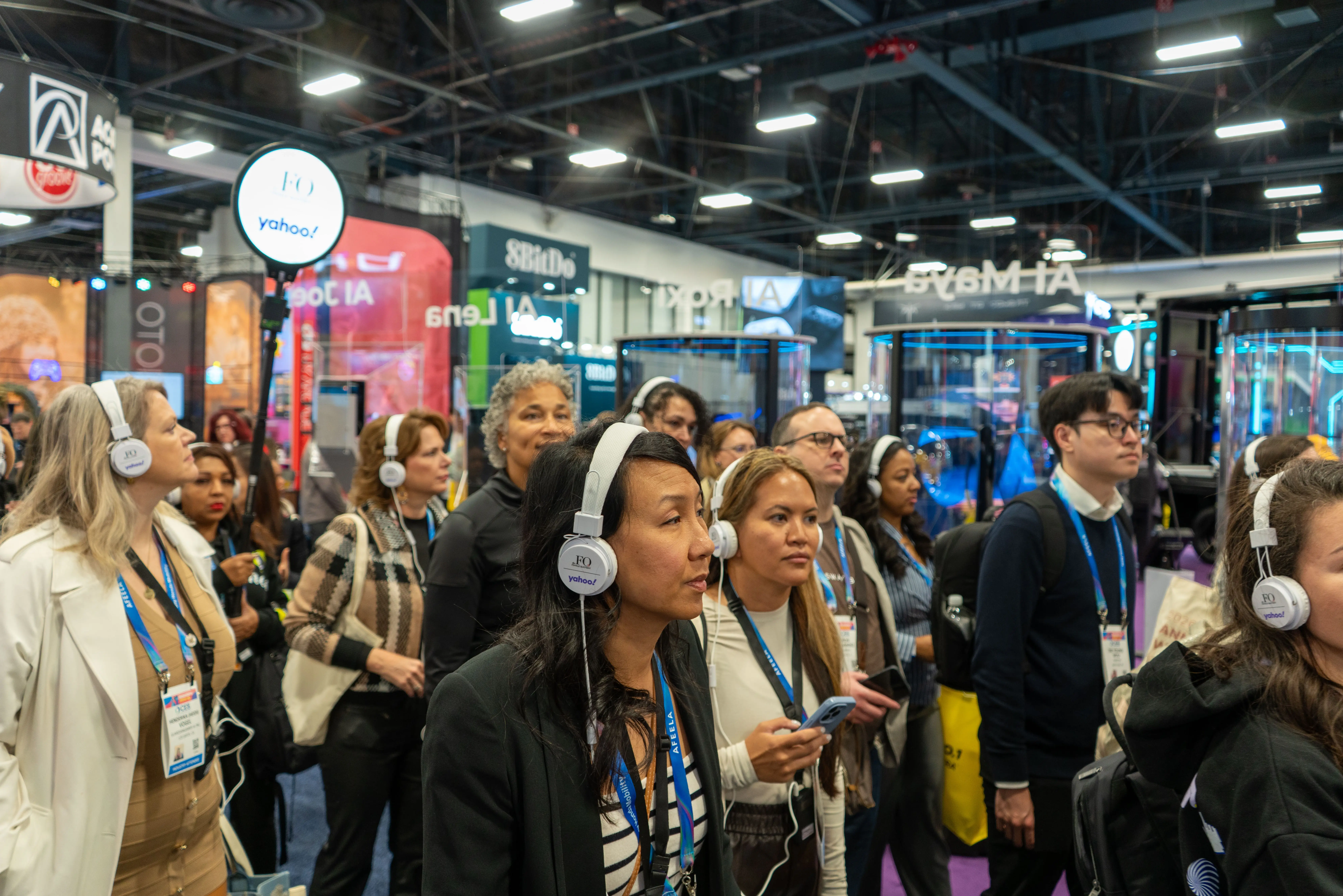 Crowd of trade show attendees wearing wireless tour guide headsets, listening together on a busy expo floor with booths and signage in the background.