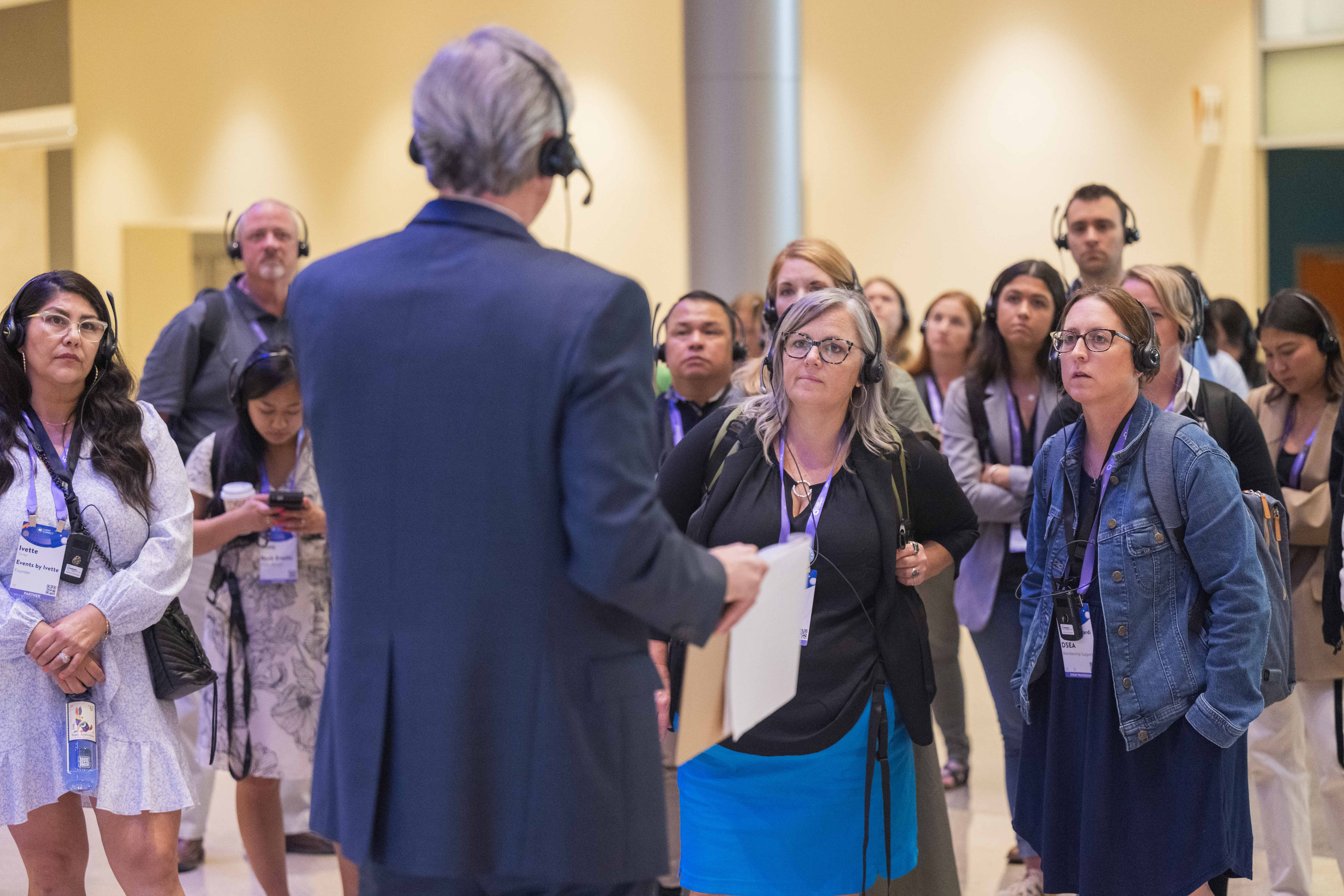 Guided tour in a convention center atrium, with an older man speaking to a group of attendees wearing headsets and event badges.